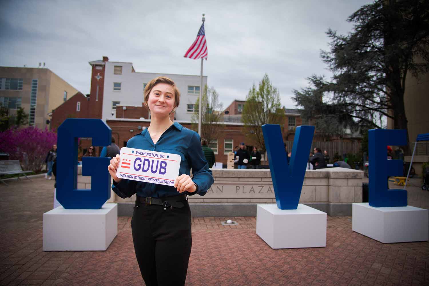 A person poses in Kogan Plaza in front of a "GIVE" statue with a DC license plate that says "GDUB"